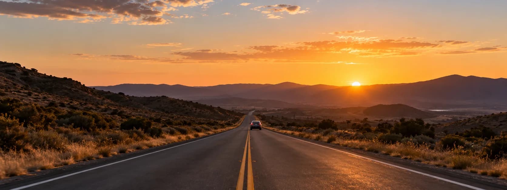 An empty two-lane highway running toward distant hills at sunset, a single car far down the road.